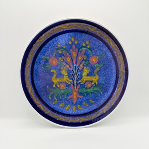 May include: Round ceramic plate with a blue center and a decorative border. The center features a colorful floral design with two yellow lion-like figures. The rim has a gold and blue pattern, and the plate has a white edge.