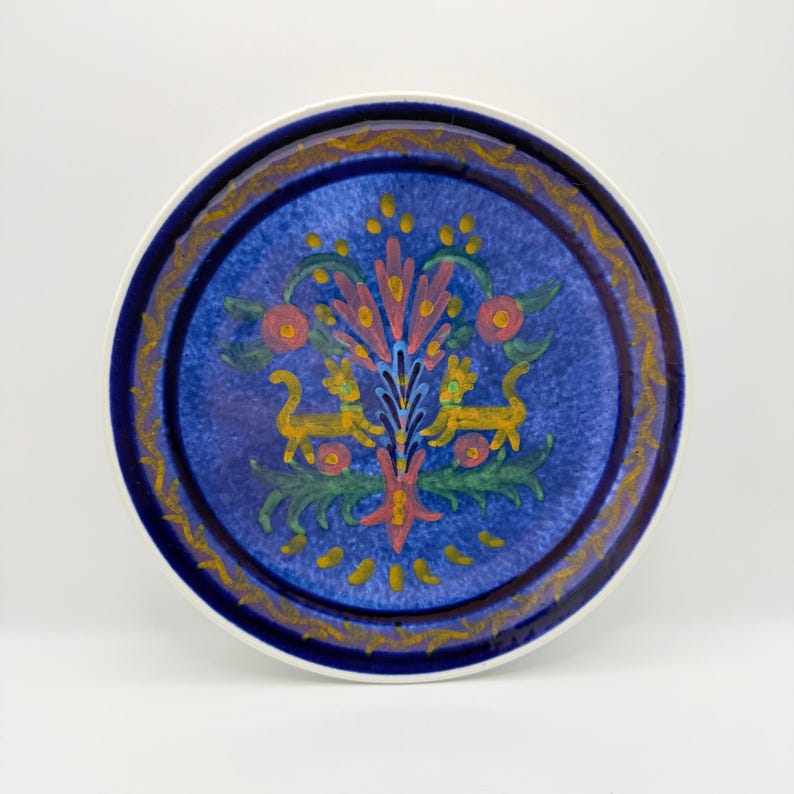 May include: Round ceramic plate with a blue center and a decorative border. The center features a colorful floral design with two yellow lion-like figures. The rim has a gold and blue pattern, and the plate has a white edge.