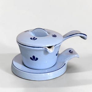 May include: A light blue enamel sauce pot with a matching lid and a small side handle, featuring a dark blue floral pattern. The pot rests on a circular base with a handle, and both pieces have dark blue trim along the edges and a spout.
