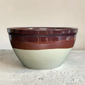 May include: A ceramic bowl featuring a two-tone design with a glossy dark brown upper section and a matte light cream lower section. The bowl has a wide, rounded rim and a smooth, curved body that tapers toward the base, showcasing a simple form.