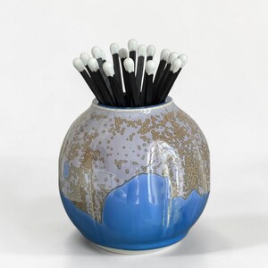 May include: A round ceramic vessel with a glossy finish featuring a blue base and a light lavender top with a speckled tan pattern. The vessel is filled with a cluster of wooden matches that have black stems and white tips, arranged upright.