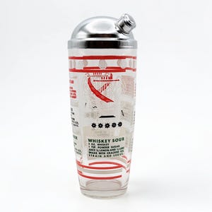 May include: A clear glass cocktail shaker with a polished chrome lid. The exterior features red and black printed text and graphics, including a recipe for a Whiskey Sour that lists 2 oz. whiskey, 1 tsp. powder sugar, and lemon and lime juice.