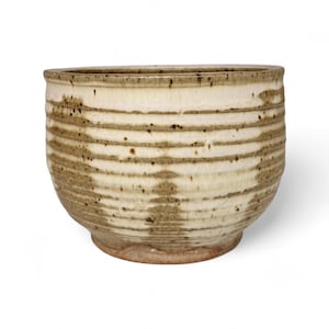 May include: A ceramic bowl with a cream-colored exterior and horizontal bands of brown glaze. The bowl has a rounded shape with a slightly flared rim and a textured surface. The bowl is likely handmade and suitable for serving food.