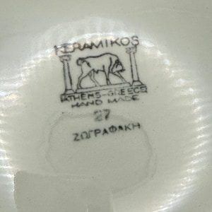 May include: Close-up of a white ceramic surface with a black and white emblem. The emblem features a drawing of an animal between two pillars, with text that reads "KERAMIKOS", "ATHENS-GREECE HAND MADE", and "27 Z&Omega;&Gamma;&Rho;&Alpha;&Phi;&Alpha;&Kappa;&Eta;".