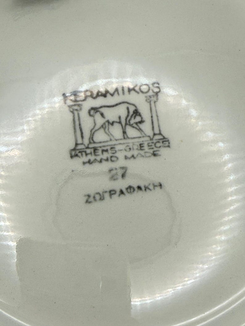 May include: Close-up of a white ceramic surface with a black and white emblem. The emblem features a drawing of an animal between two pillars, with text that reads "KERAMIKOS", "ATHENS-GREECE HAND MADE", and "27 Z&Omega;&Gamma;&Rho;&Alpha;&Phi;&Alpha;&Kappa;&Eta;".