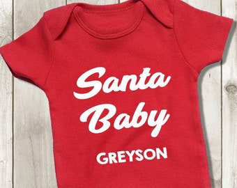 May include: A red baby bodysuit featuring the text "Santa Baby" in white script, with the name "Greyson" below. Two Christmas tree biscuits with green icing and red and white decorations are at the bottom.