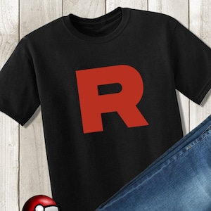 May include: Black t-shirt with a large red "R" printed on the front. A pair of blue jeans and a red and white Pokeball are also in the image.