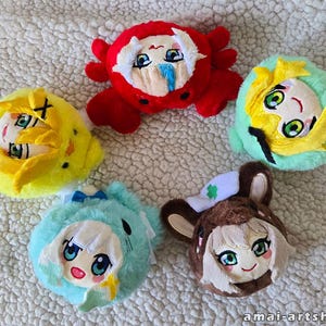 May include: Five round plush toys featuring anime-style faces. One is a red crab, one is a yellow chick, one is a light green creature, one is a light blue creature with white hair, and one is a brown animal with a white hat. Text reads amai-artshop.