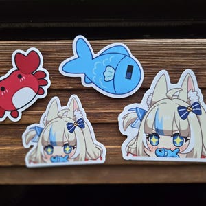 Puede incluir: Un conjunto de pegatinas con ilustraciones de dibujos animados. Incluye un cangrejo rojo, un pez azul y dos personajes de estilo anime con pelo rubio, lazos azules y accesorios de pez. Sobre una superficie de madera.