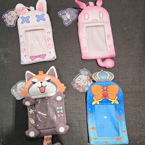 May include: Four plush card holders with clear plastic windows and attached acrylic charms. One is white with rabbit ears, one is pink with cat ears, one is a brown red panda, and one is blue with a crown and bow design. Each features a unique character.
