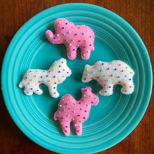 May include: Four felt animal cookies on a teal plate. The cookies are shaped like an elephant, a camel, a bear, and a dog. Each cookie is covered in colorful sprinkles.