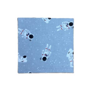 Puede incluir: Una pieza cuadrada de tela azul claro con un patr&oacute;n repetitivo de astronautas blancos en trajes espaciales con cascos negros y detalles rojos, intercalados con peque&ntilde;as estrellas blancas dispersas por toda la superficie del material.