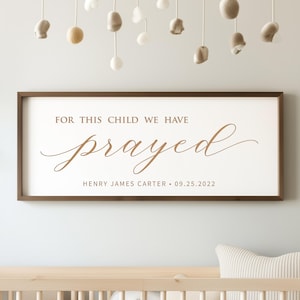 May include: A framed wooden sign with the text "FOR THIS CHILD WE HAVE prayed" in a stylish font. Below the word "prayed" is the name "HENRY JAMES CARTER • 09.25.2022". The sign is brown and white and hangs above a crib.