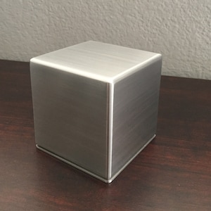 May include: A silver metal cube box with a brushed finish. The box is sitting on a dark wood surface.