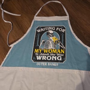 May include: A light blue and white apron with a humorous graphic. The graphic features a skeleton holding a beer and the text "WAITING FOR MY WOMAN To Admit She Was WRONG OUTER BANKS."
