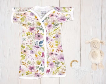 May include: A white baby gown with a floral print in shades of pink, purple, and green. The gown has a white trim and a wrap-style closure. The gown is laid out on a white wooden surface with a stuffed lamb and a wooden ring toy.