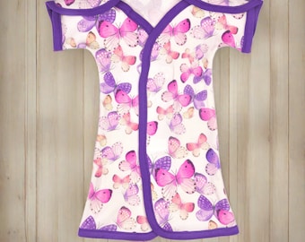 May include: A white baby gown with a purple trim and a butterfly print. The gown has short sleeves and a wrap-style closure.