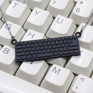 Pode incluir: Um colar de teclado preto em um teclado de computador branco. O colar é feito de um teclado impresso em 3D.