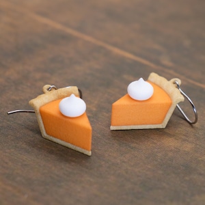 May include: A pair of earrings shaped like slices of pumpkin pie. Each earring features an orange filling, a tan crust, and a dollop of white whipped cream. The earrings hang from silver-colored hooks.
