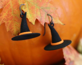 May include: A pair of dangle earrings shaped like black witch hats with orange bands. The earrings feature dark metal hooks and are positioned against a large orange pumpkin and several colorful autumn maple leaves in shades of red and yellow.