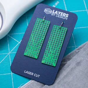 K&ouml;nnte beinhalten: Gr&uuml;ne, lasergeschnittene Ohrringe mit geometrischem Design, pr&auml;sentiert auf einer marineblauen Karte mit dem Text "Layers by Design" und "Laser Cut". Die Ohrringe sind rechteckig mit einem Gittermuster.