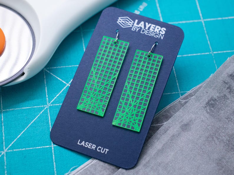 K&ouml;nnte beinhalten: Gr&uuml;ne, lasergeschnittene Ohrringe mit geometrischem Design, pr&auml;sentiert auf einer marineblauen Karte mit dem Text "Layers by Design" und "Laser Cut". Die Ohrringe sind rechteckig mit einem Gittermuster.