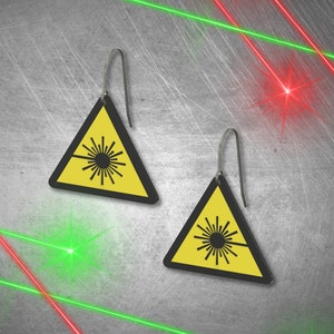 May include: A pair of laser warning sign earrings. The earrings are yellow with black borders and feature a laser beam symbol. The earrings are hanging from silver hooks.