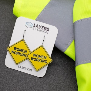 K&ouml;nnte beinhalten: Ein Paar gelbe und schwarze Ohrringe in Form eines Bauschildes mit dem Text "Women Working" auf jedem Ohrring. Die Ohrringe sind lasergeschnitten und werden auf einer wei&szlig;en Karte mit dem Text "Layers by Design" und "Laser Cut" pr&auml;sentiert.