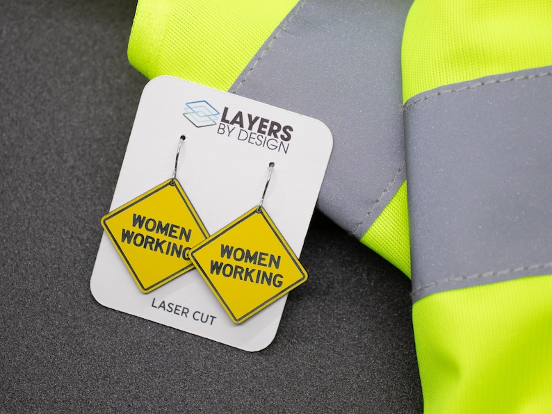 K&ouml;nnte beinhalten: Ein Paar gelbe und schwarze Ohrringe in Form eines Bauschildes mit dem Text "Women Working" auf jedem Ohrring. Die Ohrringe sind lasergeschnitten und werden auf einer wei&szlig;en Karte mit dem Text "Layers by Design" und "Laser Cut" pr&auml;sentiert.