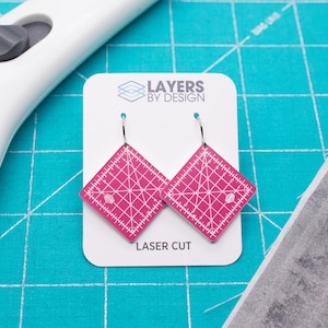 May include: A pair of diamond-shaped earrings featuring a bright pink surface with white grid lines and measurement markings resembling a quilting ruler. The earrings are attached to a white display card labeled Layers by Design and Laser Cut.
