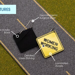 K&ouml;nnte beinhalten: Ein gelbes Schild mit dem Text "WOMEN WORKING" darauf. Das Schild ist aus laminiertem Acryl und hat einen schwarzen Metallring. Das Schild h&auml;ngt an einer schwarzen Metallkette. Das Schild befindet sich auf einer grauen Oberfl&auml;che mit einer gelben Linie.