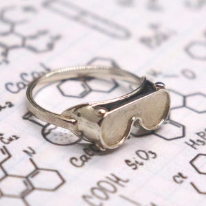 Peut inclure: Une bague en argent avec un design de lunettes miniatures. La bague est posée sur un fond de schémas et de formules scientifiques, y compris des structures chimiques. Les lunettes ont une apparence détaillée et réaliste.