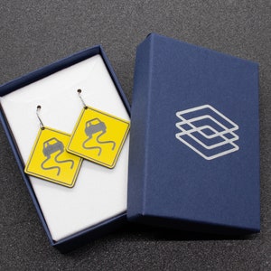 May include: A pair of earrings with yellow and black car warning signs. The earrings are in a white box with a blue lid. The box has a logo on the lid.