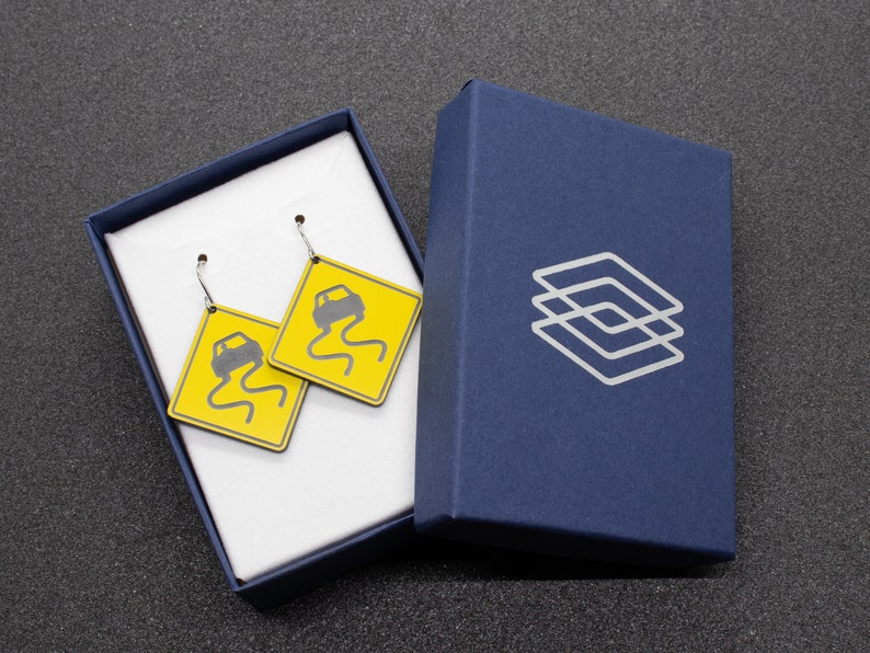 May include: A pair of earrings with yellow and black car warning signs. The earrings are in a white box with a blue lid. The box has a logo on the lid.