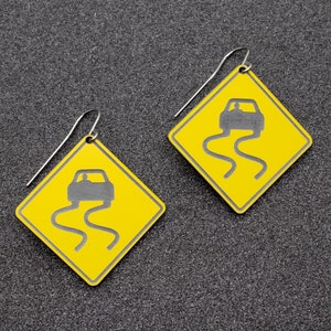 May include: A pair of yellow earrings shaped like a diamond with a black silhouette of a car driving on a winding road. The earrings are made of metal and have a silver hook.