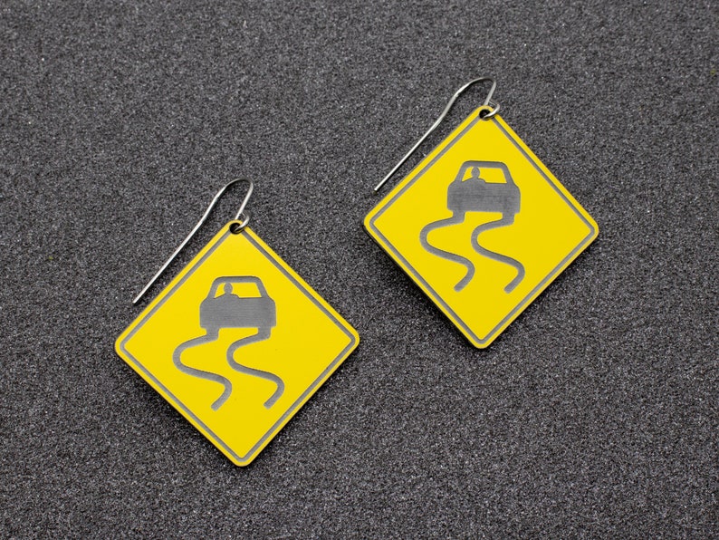 May include: A pair of yellow earrings shaped like a diamond with a black silhouette of a car driving on a winding road. The earrings are made of metal and have a silver hook.