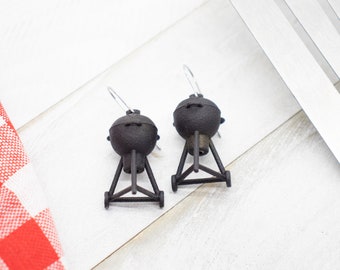 May include: A pair of dangle earrings featuring miniature black charcoal grills. Each earring is shaped like a round kettle grill with a tripod base and small wheels, suspended from a silver-toned metal hook for pierced ears.