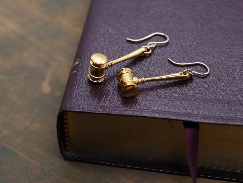 Puede incluir: Un par de pendientes de oro en forma de martillo de juez sobre un libro morado. Los pendientes tienen forma de martillo con un mango dorado y una cabeza dorada.