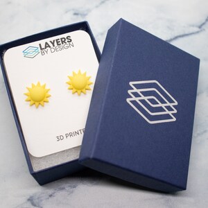 May include: A pair of yellow sun earrings in a blue box with the text "Layers by Design" and "3D Printed".