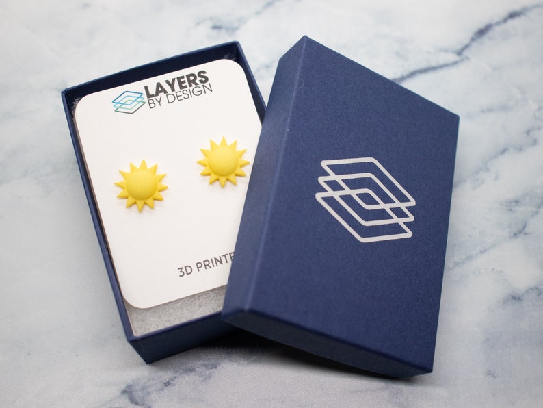 May include: A pair of yellow sun earrings in a blue box with the text "Layers by Design" and "3D Printed".