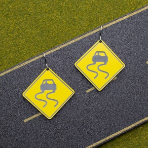 May include: A pair of yellow and black earrings shaped like road signs with a car driving on a slippery road. The signs have the text "Slippery When Wet" on them.