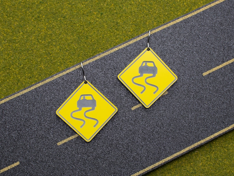 May include: A pair of yellow and black earrings shaped like road signs with a car driving on a slippery road. The signs have the text "Slippery When Wet" on them.