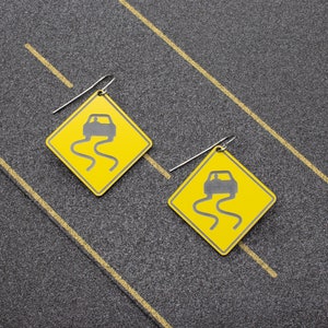 May include: A pair of earrings featuring yellow road signs with a black car icon and a black wavy line, symbolizing a slippery road. The earrings are made of metal and have a silver finish.