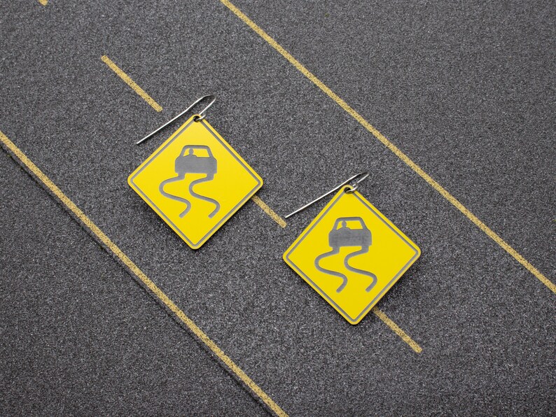 May include: A pair of earrings featuring yellow road signs with a black car icon and a black wavy line, symbolizing a slippery road. The earrings are made of metal and have a silver finish.