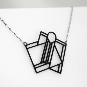Op de afbeelding: Een zwarte ketting met een geometrische hanger. De hanger is gemaakt van dun metaal en heeft de vorm van een gevouwen stuk papier.