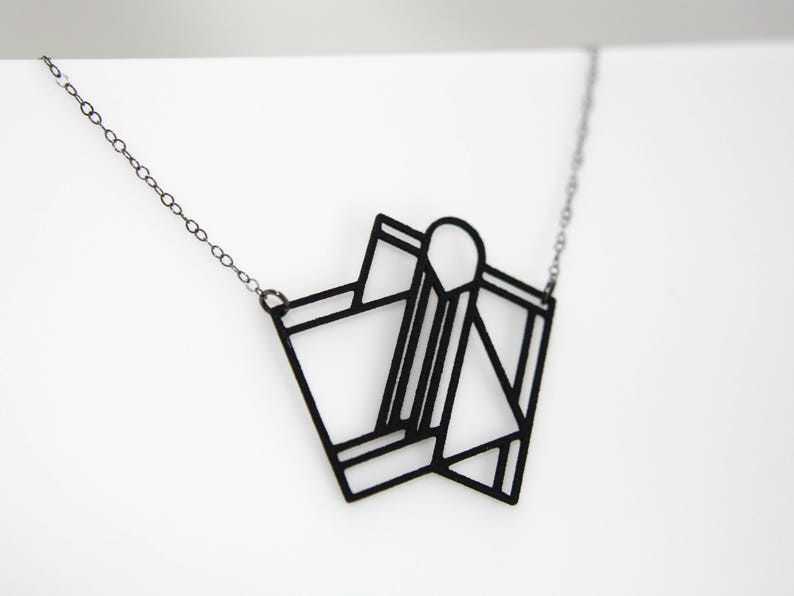 Op de afbeelding: Een zwarte ketting met een geometrische hanger. De hanger is gemaakt van dun metaal en heeft de vorm van een gevouwen stuk papier.