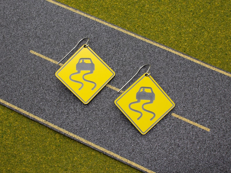 May include: A pair of yellow and silver earrings shaped like a road sign with a car driving on a slippery road. The sign reads "Slippery When Wet".