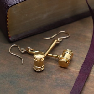 Puede incluir: Un par de pendientes de tono dorado con forma de martillo en miniatura, s&iacute;mbolo de la justicia. Los pendientes cuelgan de un libro con una cubierta de color burdeos.