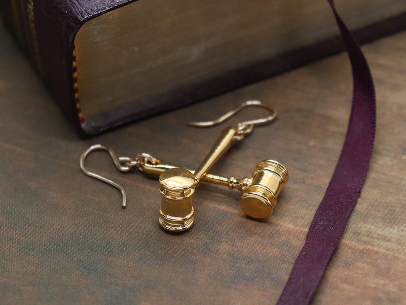 Puede incluir: Un par de pendientes de tono dorado con forma de martillo en miniatura, s&iacute;mbolo de la justicia. Los pendientes cuelgan de un libro con una cubierta de color burdeos.