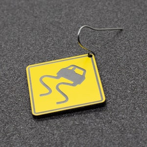 May include: A yellow and black car warning sign earring. The sign depicts a car skidding on a slippery road.